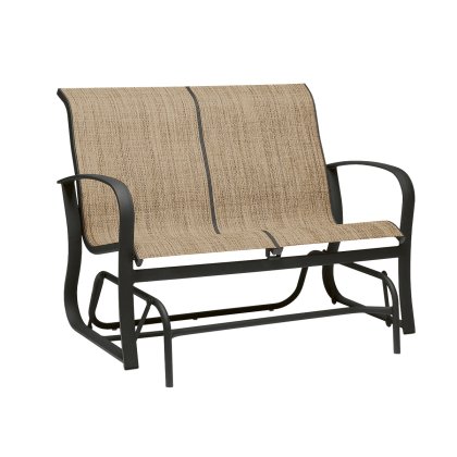 The Fremont Sling Gliding Love Seat is a two-seater outdoor bench with a metal frame and beige fabric seats. It features curved armrests, a gliding base for rocking motion, and textured woven fabric.