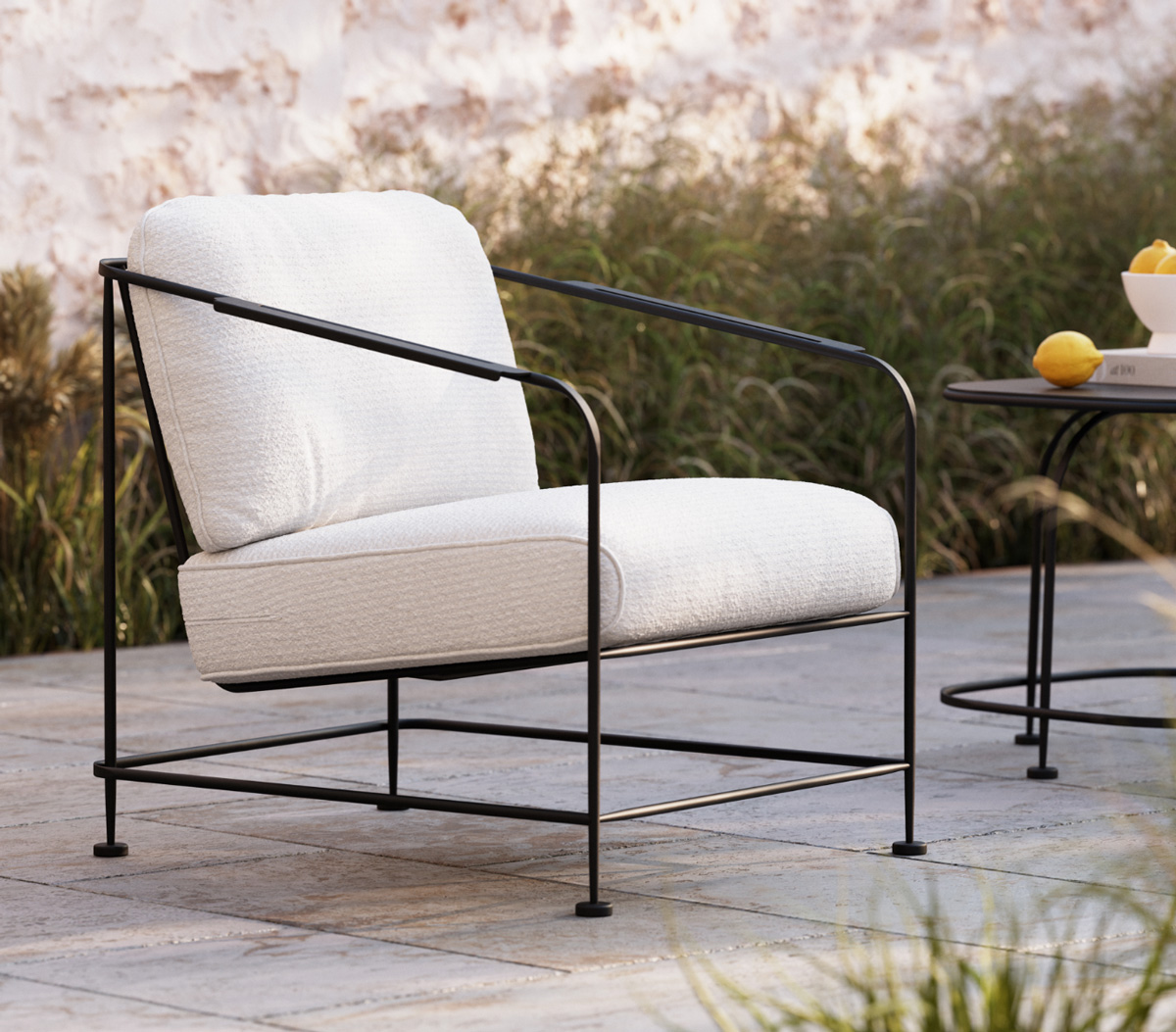 A Reed Collection modern outdoor lounge chair with a thin black iron frame and plush white cushions sits on a stone patio near a small table with lemons, surrounded by greenery and a textured wall.
