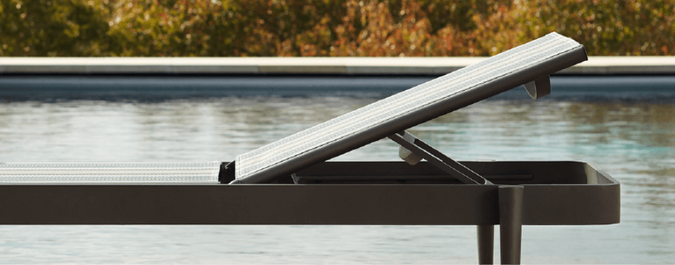 A close-up of a modern aluminum outdoor lounge chair positioned beside a swimming pool, with the backrest partially raised and water in the background.