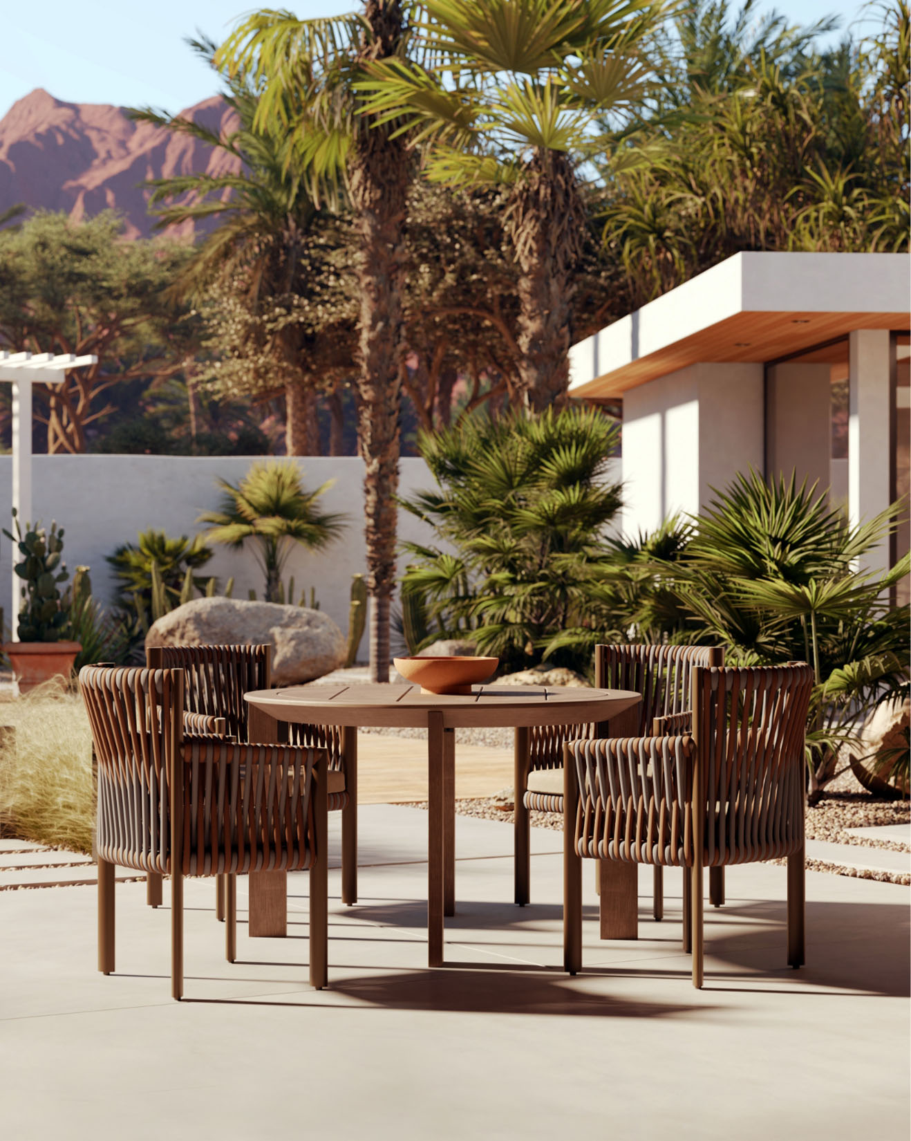 A modern patio features woven outdoor furniture: four stylish chairs and a wooden table, surrounded by palm trees and desert plants, with a contemporary house and mountains in the background.
