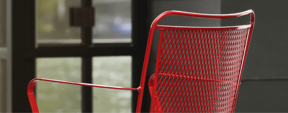 A close-up of a red iron outdoor furniture chair with a mesh backrest and armrests, set outdoors near a window with blurred glass panes in the background.