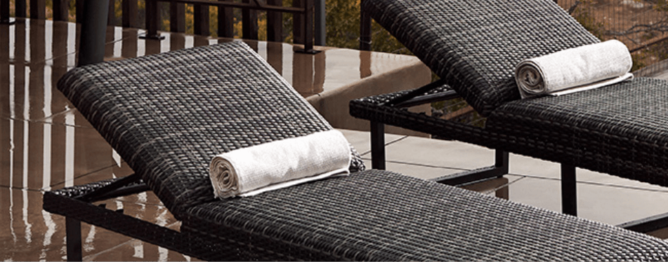 Two black woven outdoor lounge chairs with rolled white towels on the headrests are positioned outdoors on a shiny, tiled surface near a railing.